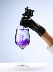 Wine glass and purple ink. Tattooer master's hand in black glove holding machine for making tattoo art on body isolated on white background. Concept of art, job, hobby, fashion, style