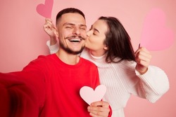 Portrait of lovely young couple, woman kissing happy and smiling man, taking selfie together isolated over pink background. Concept of love, relationship, Valentine's Day, emotions, lifestyle