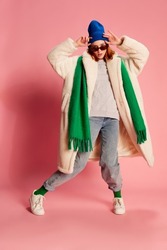 Portrait of young girl in sunglasses, blue hat, green scarf and fur coat posing over pink background. Stylish youth. Concept of beauty, winter fashion, lifestyle, emotions, facial expression. Ad