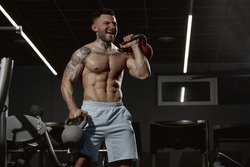 Portrait of young muscular man training shirtless in gym indoors. Hand exercises with kettlebell. Relief body shape. Concept of health, sportive lifestyle, fitness, body care, diet, strength