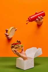 Food pop art photography. Female hand sticking out orange paper with ketchup over burger on hand sticking out food box. Concept of taste, creativity, art. Complementary colors. Copy space for ad, text