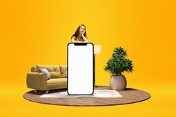 Young calm girl standing behind giant 3D model of mobile phone with empty screen for text, ad over yellow background with home interior. Online shopping, comfort. Mockup for design, logo