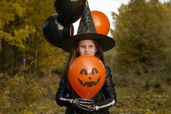 Funny baby girl in a witch costume for Halloween with black and orange balloons on the background of an autumn forest. Happy Halloween.