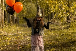 Funny baby girl in a witch costume for Halloween with black and orange balloons on the background of an autumn forest. Happy Halloween.