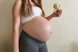 A girl pregnant at the ninth month holds a knitted rattle in the form of a lion in her hands on a monochrome light background. An impersonal photo without a head.