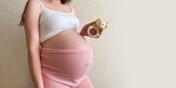 A girl pregnant at the ninth month holds a knitted rattle in the form of a lion in her hands on a monochrome light background. An impersonal photo without a head. banner.