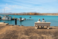 A couple of people sitting on a stone bench by a sandy beach in an ocean bay in Fuerteventura on a sunny day, hotels on a hill in the distance