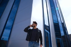 Handsome young man with beard, sculpted body and sunglasses is talking on the phone in front of a glass building. The man is wearing jeans and a leather jacket.