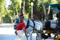 Young black woman dressed as a flamenco gypsy in a famous square in Seville, Spain. She is wearing a beige dress with ruffles and a red shawl and is standing next to a horse-drawn carriage