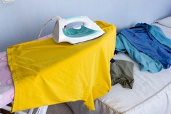 Ironing clothes on a yellow T-shirt on the ironing board and in the background more clothes to be ironed on the bed. Concept of housework.