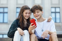 young guy and a girl sitting on the street, who have one pair of headphones for two, look into a red smartphone with a smile.