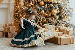 A little girl in a smart, beautiful green dress sits by the Christmas tree and opens boxes of Christmas presents