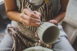 
In the image female hands trace a striped pattern to a clay mug. For this a special tool is used. Image with a selective focus.