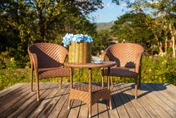 Two armchairs, wooden garden furniture on wood floor  outdoor for relaxing on summer days. Garden landscape with two chairs in nature, flowers in vase and coffee cup.. Rest in park cafe. 