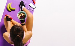Sports and nutrition. Top photo of a girl with dumbbells and muesli in hand while sitting on the mat. White background and blank advertising space.