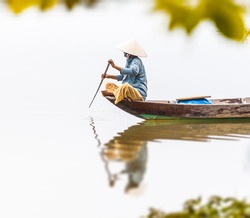 Woman in conical hat sitting on canoe and rowing. Traditional asian boat made of wood. Person holding paddle in hands. Ripple on water. Reflection of vessel in river. Travel by boat in Vietnam, Asia.