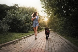 The young beautiful woman is running with the black dog in the park at sunset