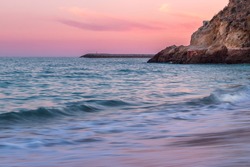 Colorful pink dreamy sunset over Atlantic Ocean coast on the beach of Albufeira city, Faro, Algarve, Portugal with soft long exposure water with limestone cliffs and lighthouse in the background
