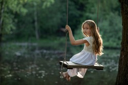 A girl in the forest swinging on a swing. Rope swing on a forest lake. Barefoot girl in a white dress with long hair. Image with selective focus.