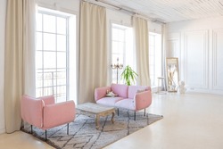 delicate and cozy light interior of the living room with modern stylish furniture of pastel pink color and white walls with stucco moldings in daylight