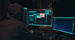 Hacker in a hood sits in front of computer screens and hacks databases. The concept of cyberterrorism, server hacking and cyberattacks. The room is dark, a person is typing code into the command line