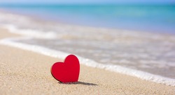 A red little heart stands on a sandy beach. In the background, a sea wave.