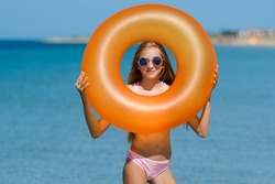 Portrait of a joyful girl on the beach with an orange rubber ring. The girl looks through the hole in the rubber ring.
