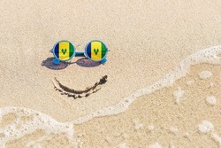 A painted smile on the sand and sunglasses with the flag of Saint Vincent and the Grenadines. The concept of a positive and successful holiday in the resort of Saint Vincent and the Grenadines.