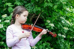 The girl plays a sad melody on the violin. A child with a violin on the background of a blossoming tree. The child learns to play the violin. Art for the kid. Little girl with a musical instrument. 