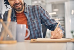 Close up of male worker in headset making notes and smiling while sitting at the table in office
