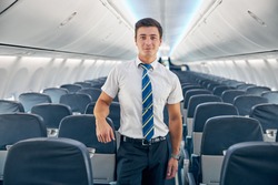 Close up portrait of handsome confident steward posing at the photo camera near the cozy chair of passenger aircraft
