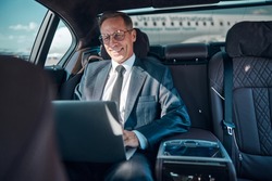 Cheerful elegant man in glasses is sitting in car with notebook while being transfered at airport