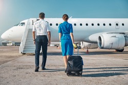 Man and woman in aviation uniform are walking to plane stairway with luggage before departure