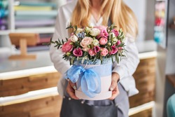 Cropped photo of a blonde lady holding a cylinder box of various flowers while standing in a floral studio