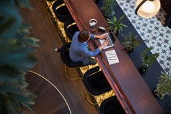 Top view photo of a focused gentleman sitting on stool while typing on laptop at a fancy hotel lounge