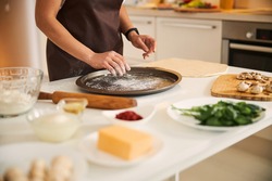 Close up photo of a woman sifting some flour on a round baking pin while cooking pizza