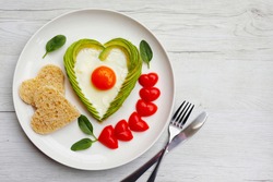 Valentine's breakfast-Fried egg in heart shaped avocado, heart shaped whole wheat bread and heart shaped tomatoes on white plate with white wood table background.Love healthy food concept.Top view