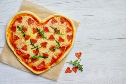 Heart shaped Italian pizza with pepperoni,arugula,pizza sauce and mozzella cheeses on parchment paper with white wood table background.Love concept for Valentine's day.Top view.Copy space

