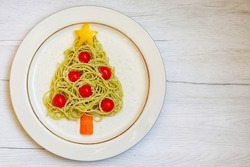 Christmas tree pesto spaghetti made it from spaghetti,pesto sauce,cherry tomato,parmesan cheeses,yellow bell pepper and carrot on plate with white wood background. Art food idea for Christmas dinner