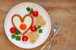 Heart shaped sausage with fried egg served with heart shaped bread and tomatoes on plate with white wood background.Romantic art food idea for Valentine's breakfast.Top view.Copy space


