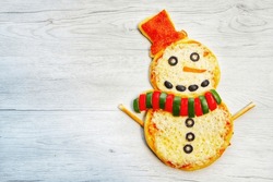 Snowman pizza made it from pizza crust,pizza sauce,mozzarella cheeses,bell peppers,black olives,pepperoni and carrot on white wood background.Creative art food idea for celebrate Christmas.Top view