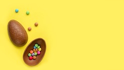 flat lay of easter chocolate egg with colorful chocolate drops inside on yellow background with copy space