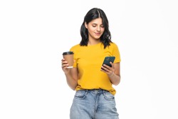 Portrait of a satisfied young woman using mobile phone while holding cup of coffee to go over white background