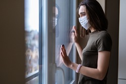Sad young woman in a medical mask looks out of the window. Quarantine during the coronavirus pandemic.