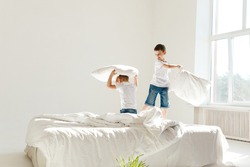Indoor positive activity. Small children jumping on a bed and having fun fighting with pillows in sunshine