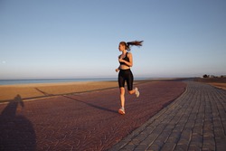 girl runner in a black tracksuit and sneakers runs by the sea during sunset