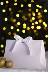 White cardboard bag with a white bow without a logo stands on a background of blurry lights. Bokeh from a golden garland and a corporate package as a gift. Beautiful cardboard bag for packing.