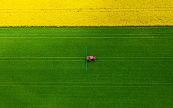 A red tractor irrigating in green grass taken from a distance from just above, tractor tracks on green grass and yellow blooming plants above