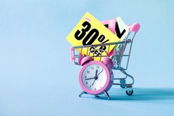 Mini shopping cart with a thirty percent discount sign and pink alarm clock on blue background. Conceptual image of sale, seasonal discounts in shopping stores, discount time	