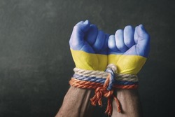 War in Ukraine. Male hands in the colors of the flag of Ukraine tied with a rope in the colors of the flag of Russia on a dark background. Conceptual image of violence, occupation, aggression